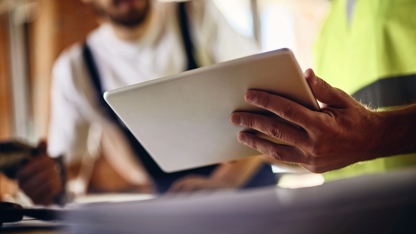Close-up: Worker's hands in a safety vest hold/review a digital tablet; second worker is blurred behind.